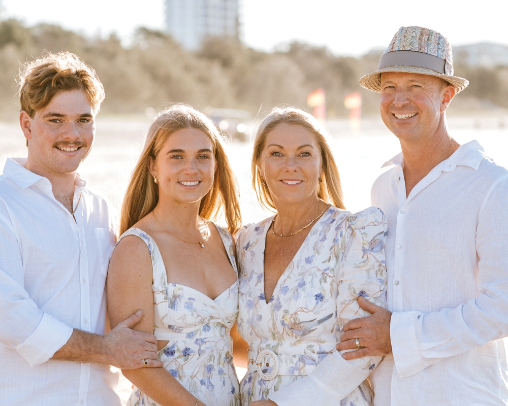 Andrew and his family smiles at the camera on a local beach. Andrew, his wife, and two children are all wearing white or light-colored clothing.