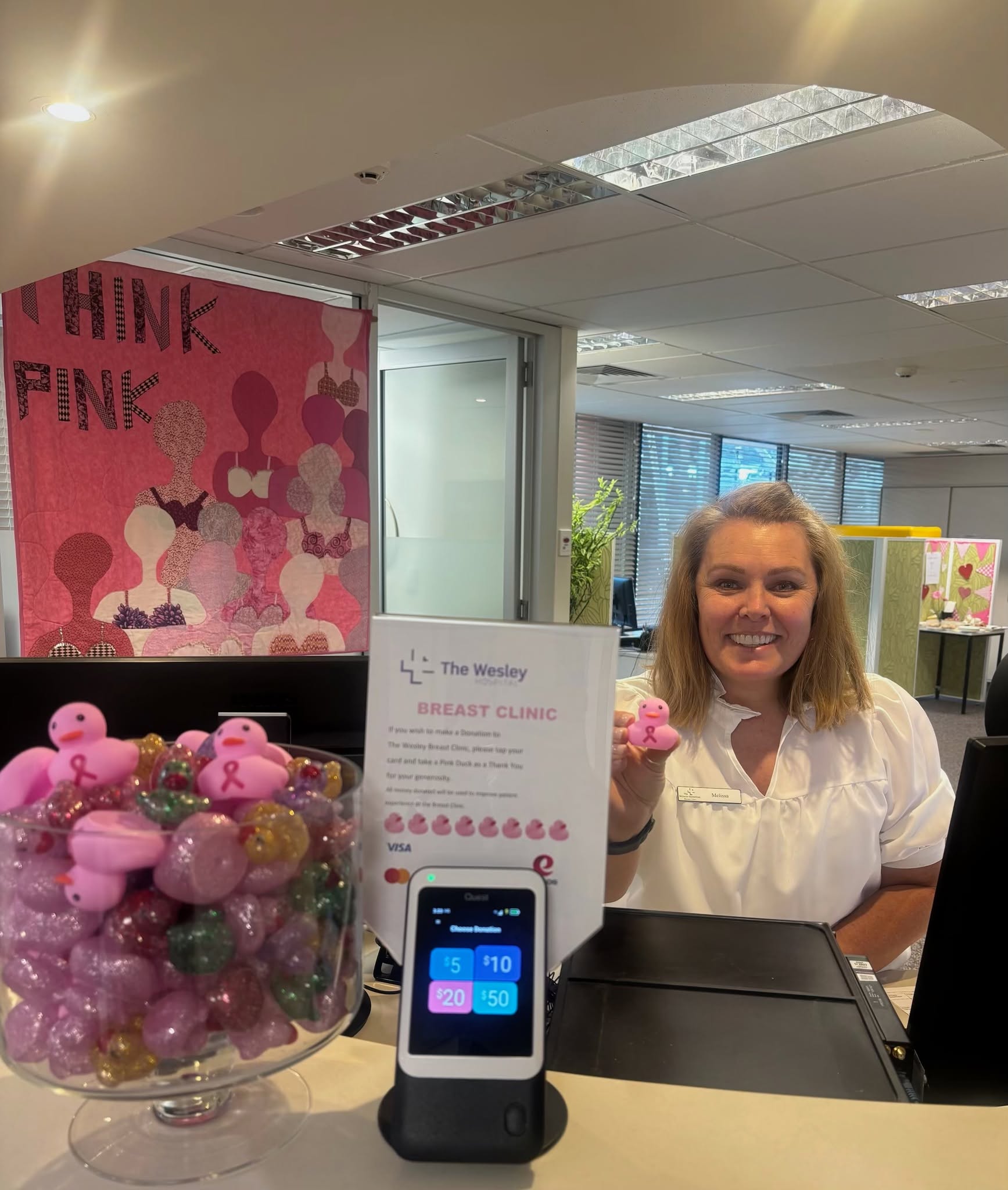 An reception desk display with a pink “THINK PINK” banner. A Wesley team member in a white shirt behind the counter is holding up a little pink duck next to a big bowl of ducks and a small tap to donate screen.