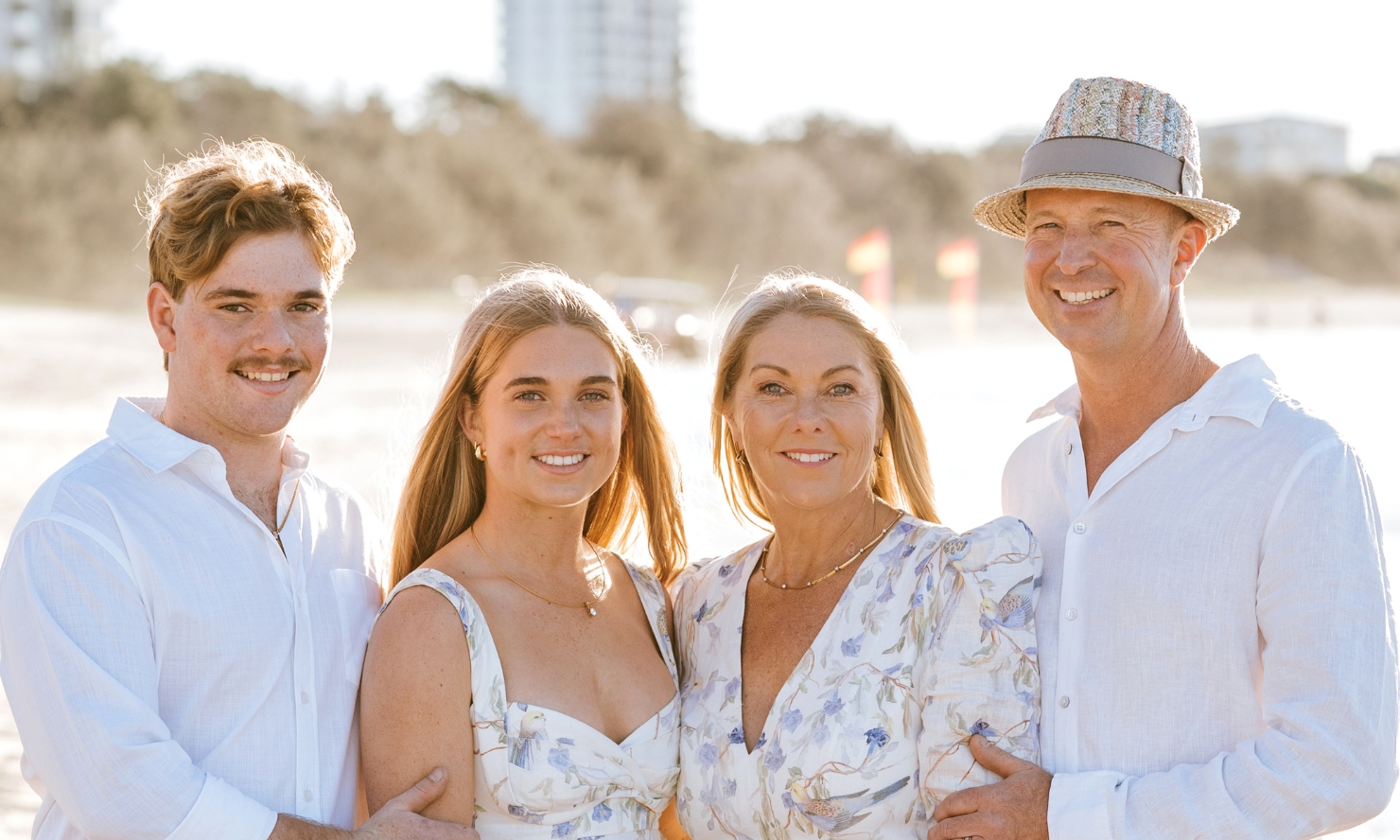 Andrew and his family smiles at the camera on a local beach. Andrew, his wife, and two children are all wearing white or light-colored clothing.