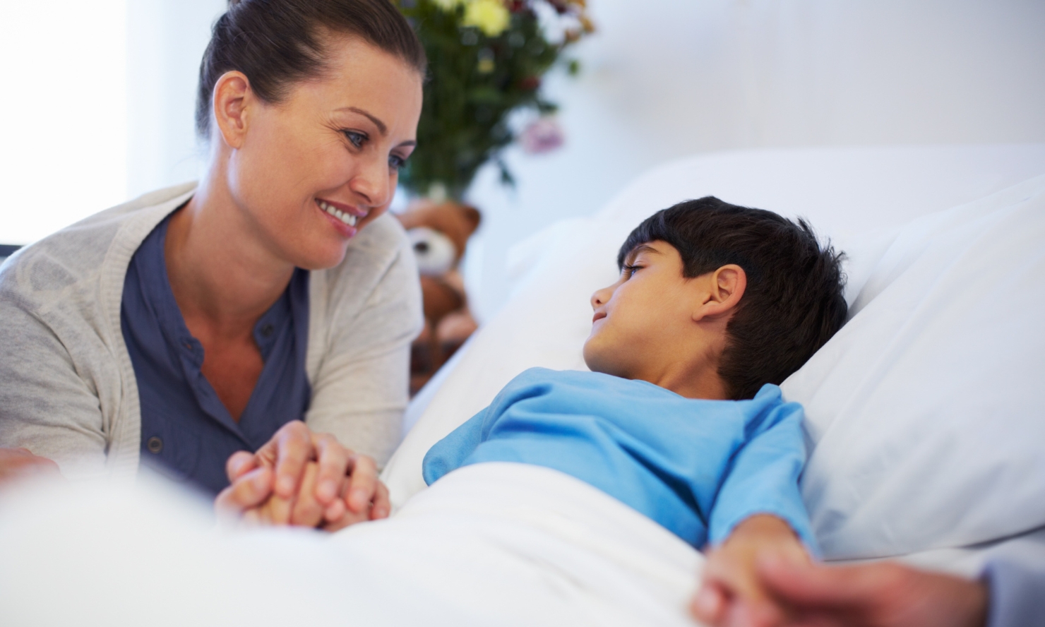 A nurse and a parent are gently holding the hands of a young boy in a hospital bed A nurse and a parent are gently holding the hands of a young boy in a hospital bed